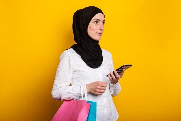 Schattige jonge moslimvrouw in hijab houdt telefoon en boodschappentassen op gele achtergrond Banner