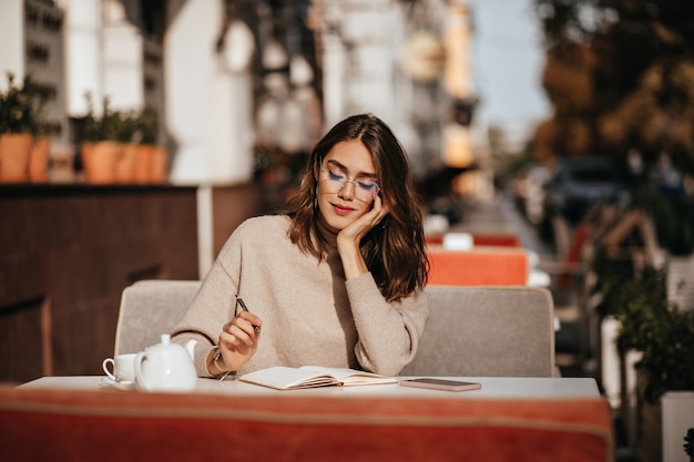 Schattige jonge student met donkerbruin golvend kapsel in beige trui en bril geconcentreerd studeren op het terras van het stadscafé op zonnige herfstdag