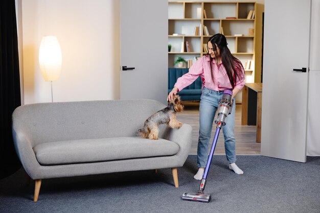 Schattige jonge vrouw stofzuigt thuis met een draadloze stofzuiger terwijl haar schattige hond toekijkt