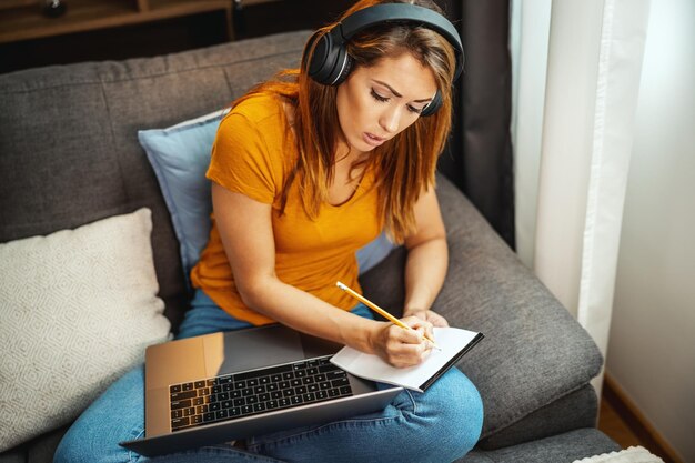 Schattige jonge vrouwelijke student zittend op de bank, met behulp van haar laptop en koptelefoon om online lessen te kijken en thuis te leren. Jonge vrouw die aantekeningen maakt terwijl ze de leraar volgt die les op video geeft