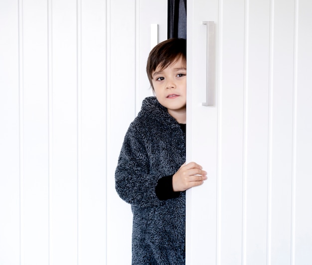 Schattige jongen draagt pluizige pyjama's die achter de garderobe staan, kleine jongen met lachend gezicht terwijl de witte deur wordt vastgehouden, Kind verstoppertje speelt en zich verstopt in de kast