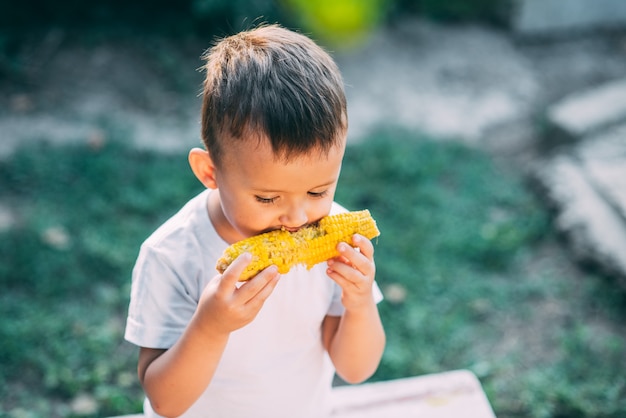 Schattige jongen in de tuin of buiten Park gekookte maïs eten is erg smakelijk in de zomer