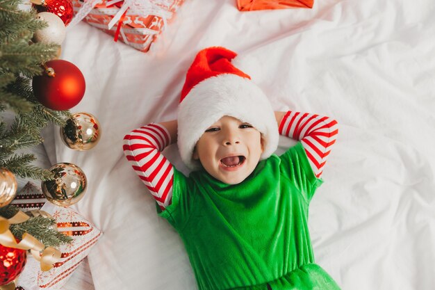 Schattige jongen in kerstman hoed ligt op een deken in de buurt van de kerstboom. gelukkig kind ligt in de buurt van de kerstboom en glimlacht