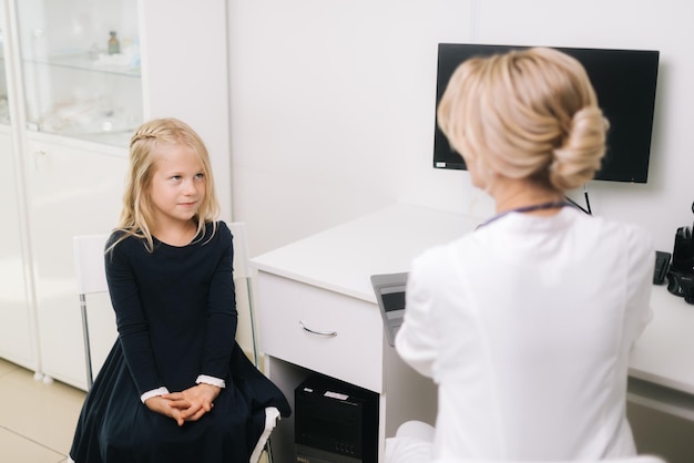 Schattige jongen meisje patiënt met blond haar zitten op stoel praten met vrouw arts. Jonge vrouw arts kinderarts in gesprek met kind meisje patiënt op medische controle afspraak