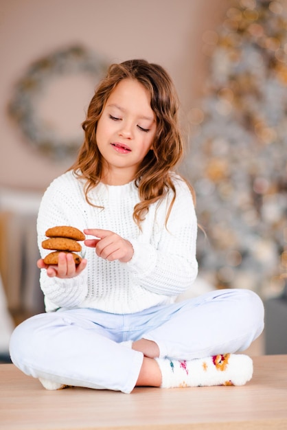 Schattige jongen meisje zelfgemaakte smakelijke koekjes eten zittend op de keukentafel over de kerstboom