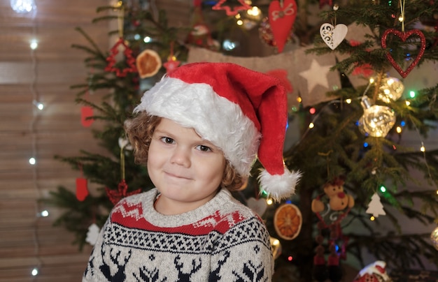 Schattige jongen met krullend haar in een rode kerstmuts bij de kerstboom. Kerstmis en Nieuwjaar concept