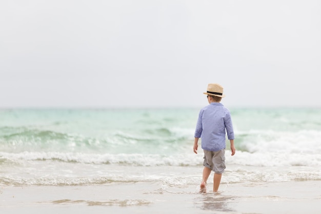 Schattige jongen met strohoed die op het strand van de oceaan loopt en met golven speelt Vakanties aan zee