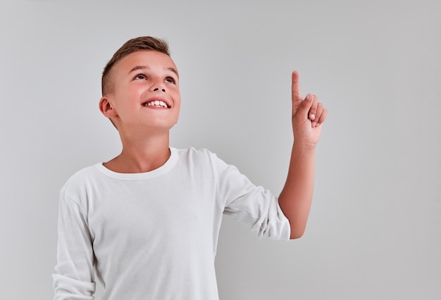 Schattige jongen op een grijze achtergrond wijst met zijn hand omhoog, kijkt op en glimlacht