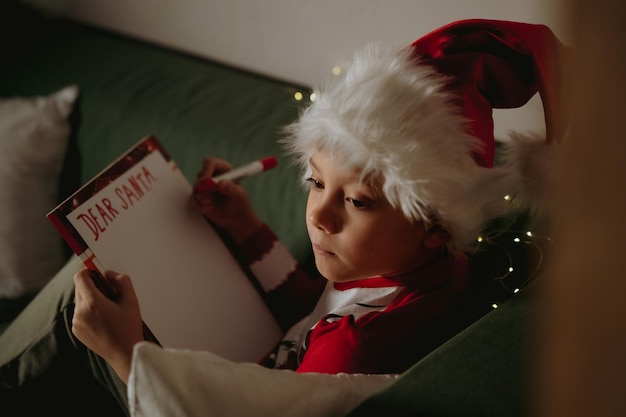 Foto schattige kaukasische kleine jongen in santa t-shirt en hatwriting brief aan de kerstman zittend op de bank. afbeelding met selectieve focus