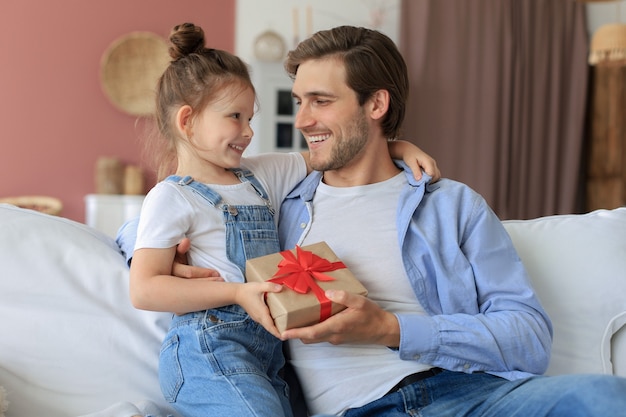 Schattige kinderdochter verrast papa, klein meisje presenteert geschenkdoos aan vader die op de bank zit. Vaderdag.