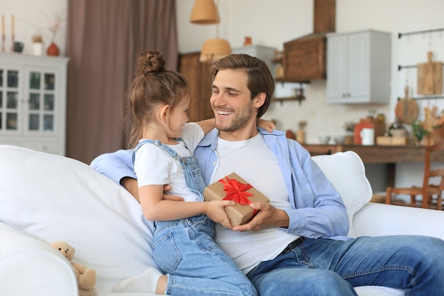 Schattige kinderdochter verrast papa, klein meisje presenteert geschenkdoos aan vader die op de bank zit. vaderdag.