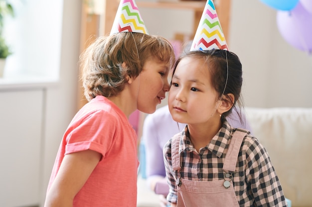 Schattige kinderen in verjaardagskapjes die een kinderachtig spel spelen waarbij je hetzelfde woord tegen elkaar moet fluisteren