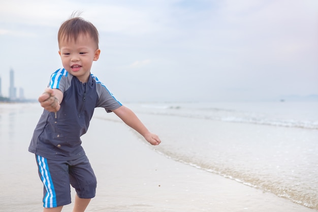 Schattige kleine Aziatische 2 jaar oude peuter baby jongenskind op strand met vuile handen bedekt met nat zand. Familie reizen, water buitenactiviteit op strandvakantie in de zomer, zintuiglijk spel met zand concept