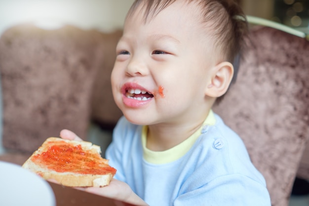 Schattige kleine Aziatische 2 jaar oude peuter babyjongen kind bijten & toast eten als ontbijt in restaurant, kind met hotel ontbijtbuffet, zelfvoeding concept, zachte & selectieve focus