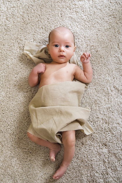 Schattige kleine baby bedekt met een stuk linnen stof ligt op het zachte tapijt