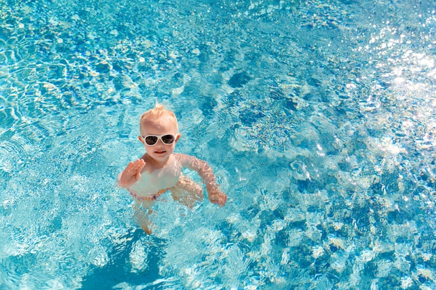 Schattige kleine baby in zonnebril zwemmen in het zwembad en zijn hand zwaaien.