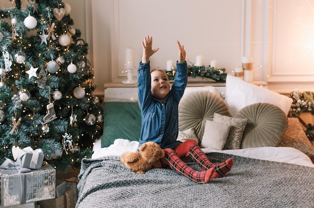 Schattige kleine baby zit met zijn handen omhoog op het bed tegen de achtergrond van de kerstboom