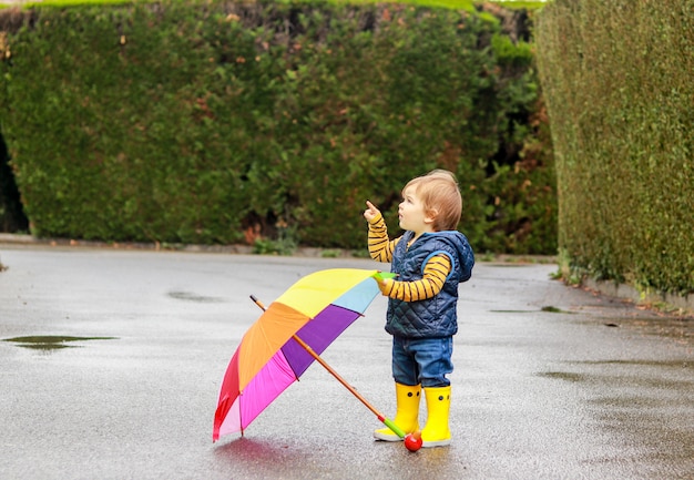 Schattige kleine babyjongen in gele rubberen laarzen met kleurrijke regenboog paraplu op natte weg achterin