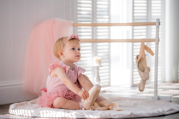 schattige kleine ballerina in een roze balletkostuum zit vlakbij de barre in de kamer en probeert haar pointe-schoenen aan te trekken Kid en balletdans Copyspace