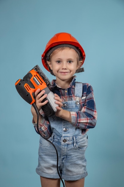 Schattige kleine blanke jongen draagt een oranje veiligheidshelm en houdt een elektrische schuurmachine bij de hand. Concept van bouw en reparatie.