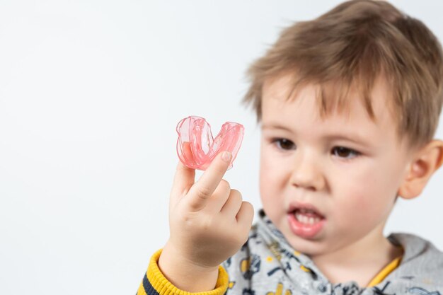 Schattige kleine blanke jongen met blond haar houdt een roze tandheelkundige myofunctionele trainer op wit op de achtergrond. tandheelkundige tariner is gemaakt om de groeiende tanden te egaliseren en de beet te corrigeren