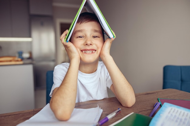 Schattige kleine blanke schooljongen met beugel die een leerboek boven zijn hoofd houdt en glimlacht