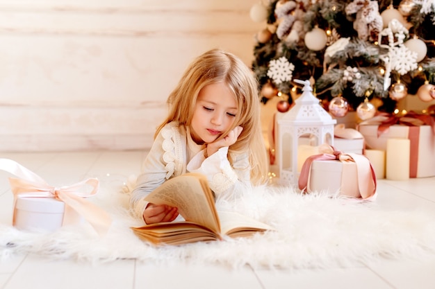 Schattige kleine blonde meisje leest thuis een boek in de buurt van de kerstboom