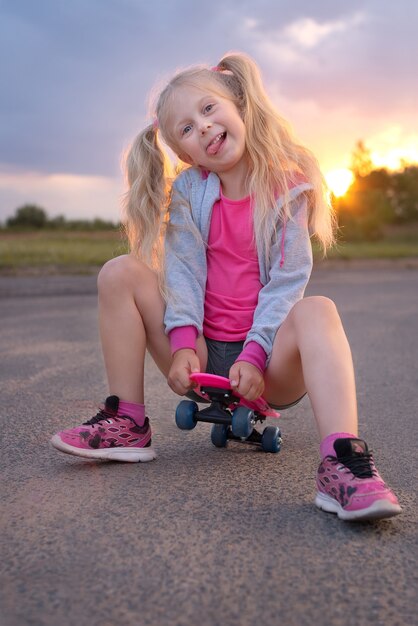 Schattige kleine blonde meisje, zittend op het roze schaatsbord