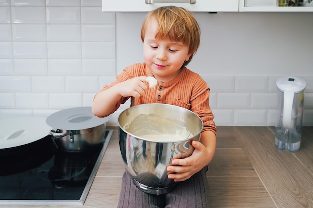 Schattige kleine blonde peuter jongen bakken taart in de binnenlandse keuken binnenshuis Glimlachend kind proeven van zoet deeg Kleine helper