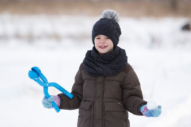 Schattige kleine jonge grappige tandeloze kind jongen in warme kleding spelen plezier maken van sneeuwballen op koude winterdag op witte heldere wazig kopie ruimte achtergrond. Buitenshuis activiteit, vakantiespelletjes.