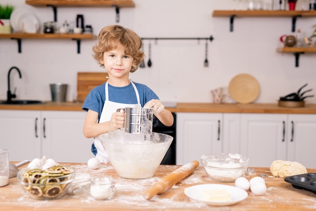 Schattige kleine jongen die bloem over de kom zift terwijl hij deeg gaat maken voor zelfgemaakt gebak in de keuken