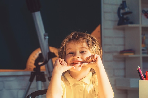 Schattige kleine jongen die naar de camera kijkt terwijl hij met de hand op de kin zit aan het bureau in de kleuterschool