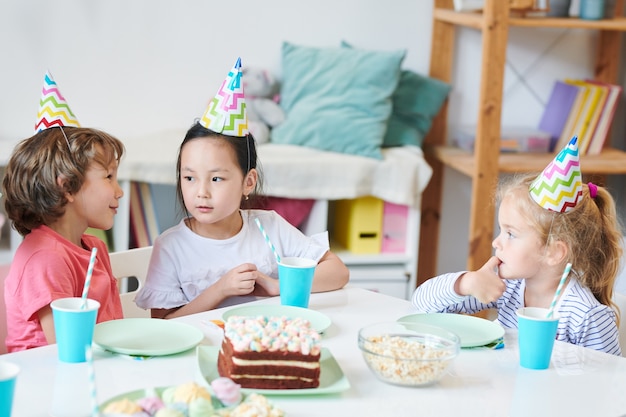 Schattige kleine jongen iets vertellen aan vrij Aziatisch meisje in verjaardag GLB door feestelijke tafel terwijl het hebben van drankjes en dessert