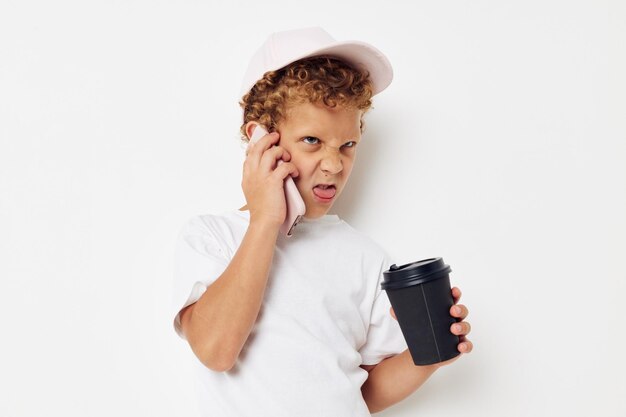 Schattige kleine jongen in een witte t-shirt pet met een telefoon in een glas met een drankje lichte achtergrond ongewijzigd