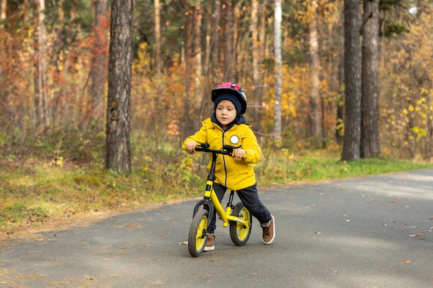 Schattige kleine jongen in gele jas en beschermende helm rijden op balansfiets langs asfaltweg in park tegen pijnbomen in het weekend