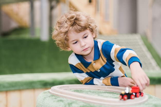 Schattige kleine jongen in vrijetijdskleding liggend op de vloer van de speeltuin tijdens het spelen van speelgoedtreinen in de kleuterschool of het recreatiecentrum