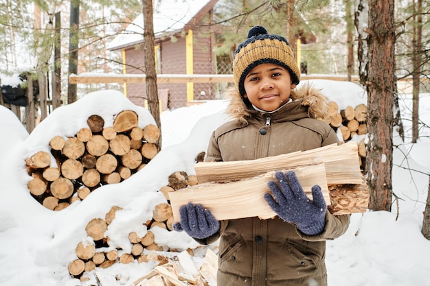 Schattige kleine jongen in warme winterjas en gebreide muts met brandhout