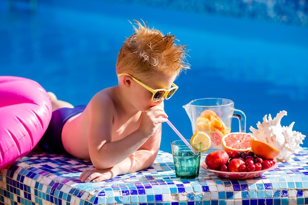 schattige kleine jongen in zonnebril en zwembroek drinkt limonade bij het zwembad.