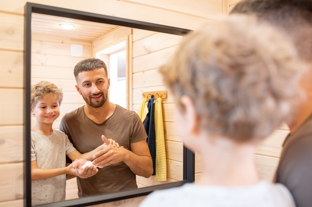 Schattige kleine jongen met blond krullend haar, kijkend naar zijn vader die schuim op het gezicht gaat aanbrengen voordat hij zich voor de spiegel in de badkamer gaat scheren