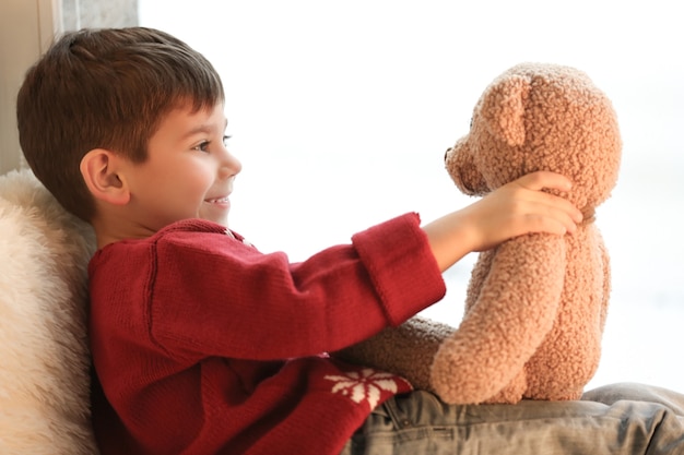 Schattige kleine jongen met teddybeer in de buurt van raam thuis