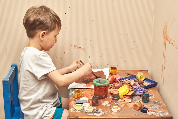 Schattige kleine jongen tekent een foto met penseel en bruine kleur aan tafel met vuile potten en speelgoed