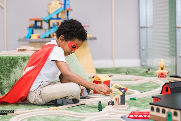 Schattige kleine jongen van afrikaanse afkomst, gekleed in witte vrijetijdskleding en rode mantel van superman spelen in de kleuterschool