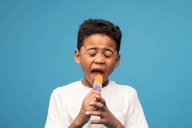 Schattige kleine jongen van Afrikaanse afkomst likken lekker ijs met smaak van sappige sinaasappel tegen blauw