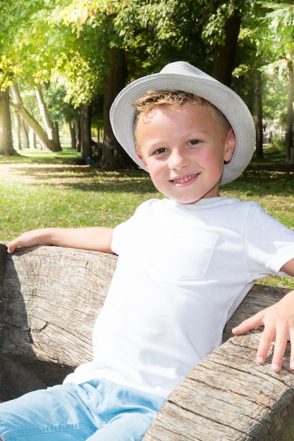 Schattige kleine jongen voor een weide zit op bank met hoed