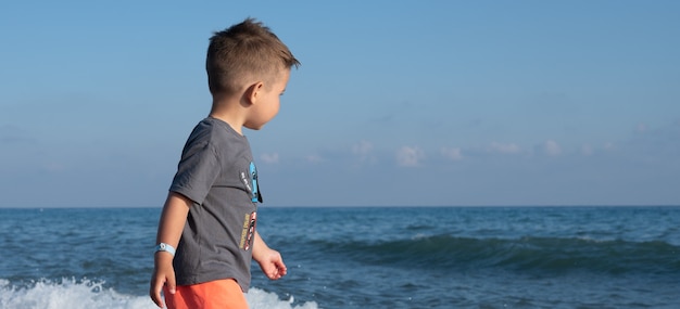 Schattige kleine jongen wandelen op het strand in schuimende golven van de zee.