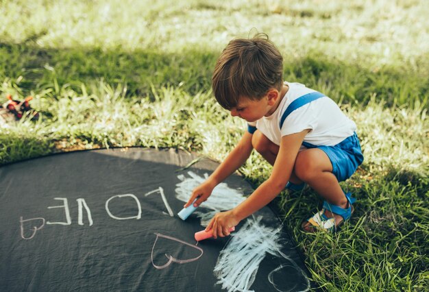 Schattige kleine jongen zittend op het groene gras spelen met kleurrijke krijtjes gelukkig kind tekenen met krijtjes in het park jeugdconcept