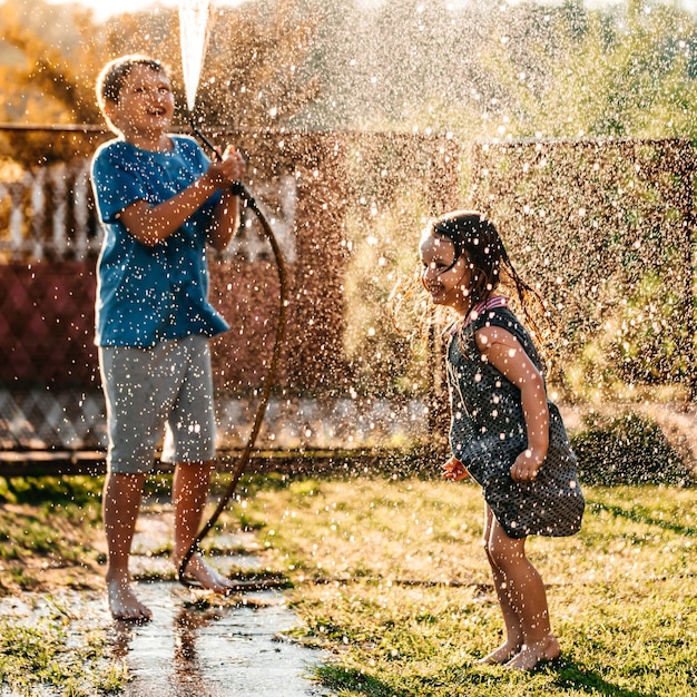 Schattige kleine kinderen spelen samen met een tuinslang op warme en zonnige zomerdag bij zonsondergang