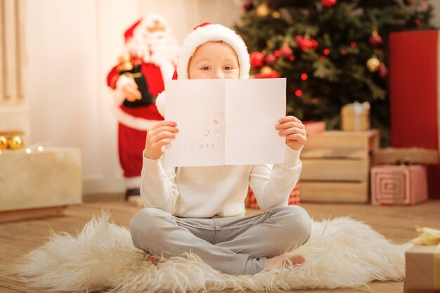 Schattige kleine kunstenaar met een kerstman hoed zittend op de vloer en verstopt zich achter zijn nieuwe meesterwerk met een kerstboom erop.