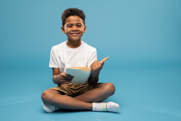 Schattige kleine schooljongen van Afrikaanse afkomst die open boek dicht bij zijn gezicht houdt en uit de omslag gluurt terwijl hij op de vloer zit