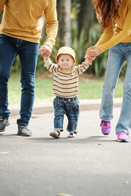 Schattige kleine Vietnamese jongen in hoed trui en spijkerbroek hand in hand van zijn ouders tijdens het wandelen in het park